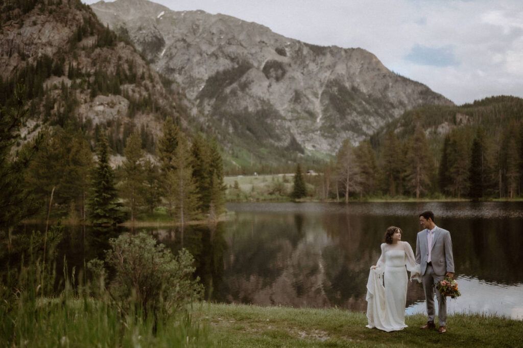 A wedding couple holding hands and standing in front of a mountain lake 