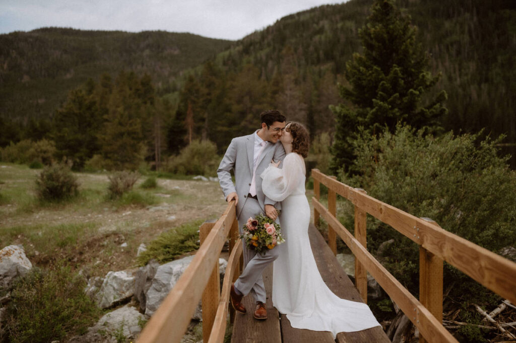 A person kissing their partner's cheek as they stand on a narrow wooden bridge in the mountains 