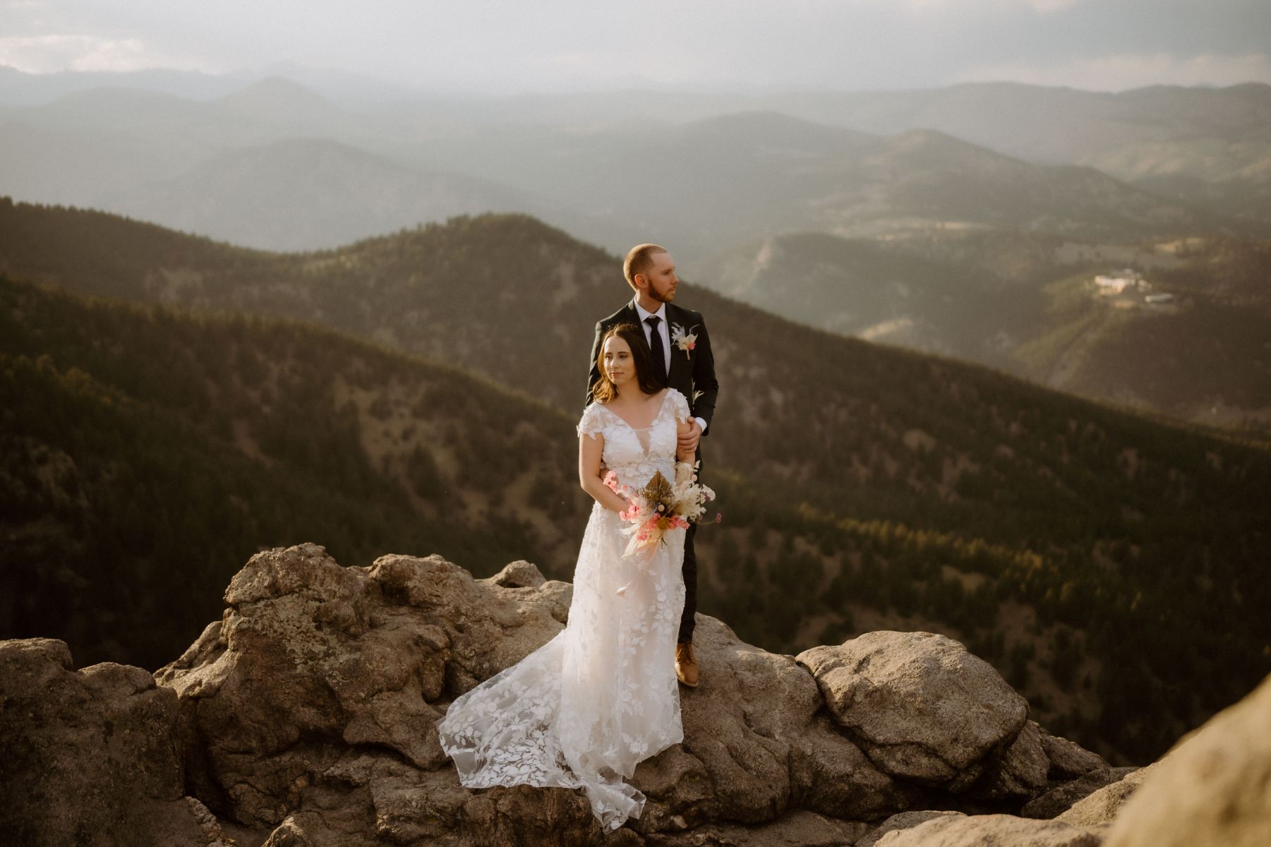 a couple standing on the overlook at lost gulch in colorado the woman is wearing a white wedding dress and holding a boquet of flowers her partner is behind her grabbing her arm and wearing a black suit 