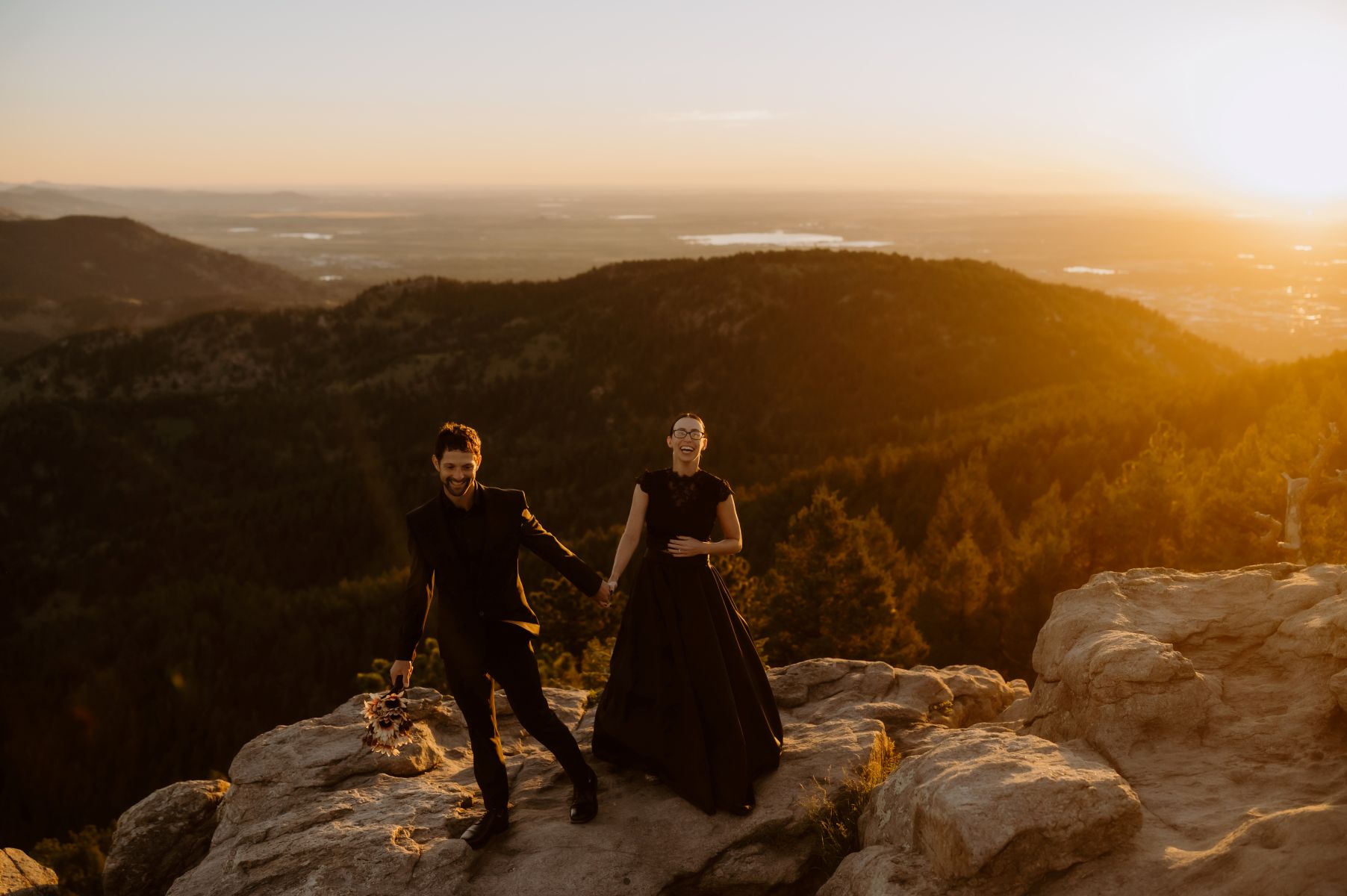a couple standing on an overlook on top of rocks holding hands and smiling and laughing at sunset the woman is wearing a black dress the man is wearing a black suit 