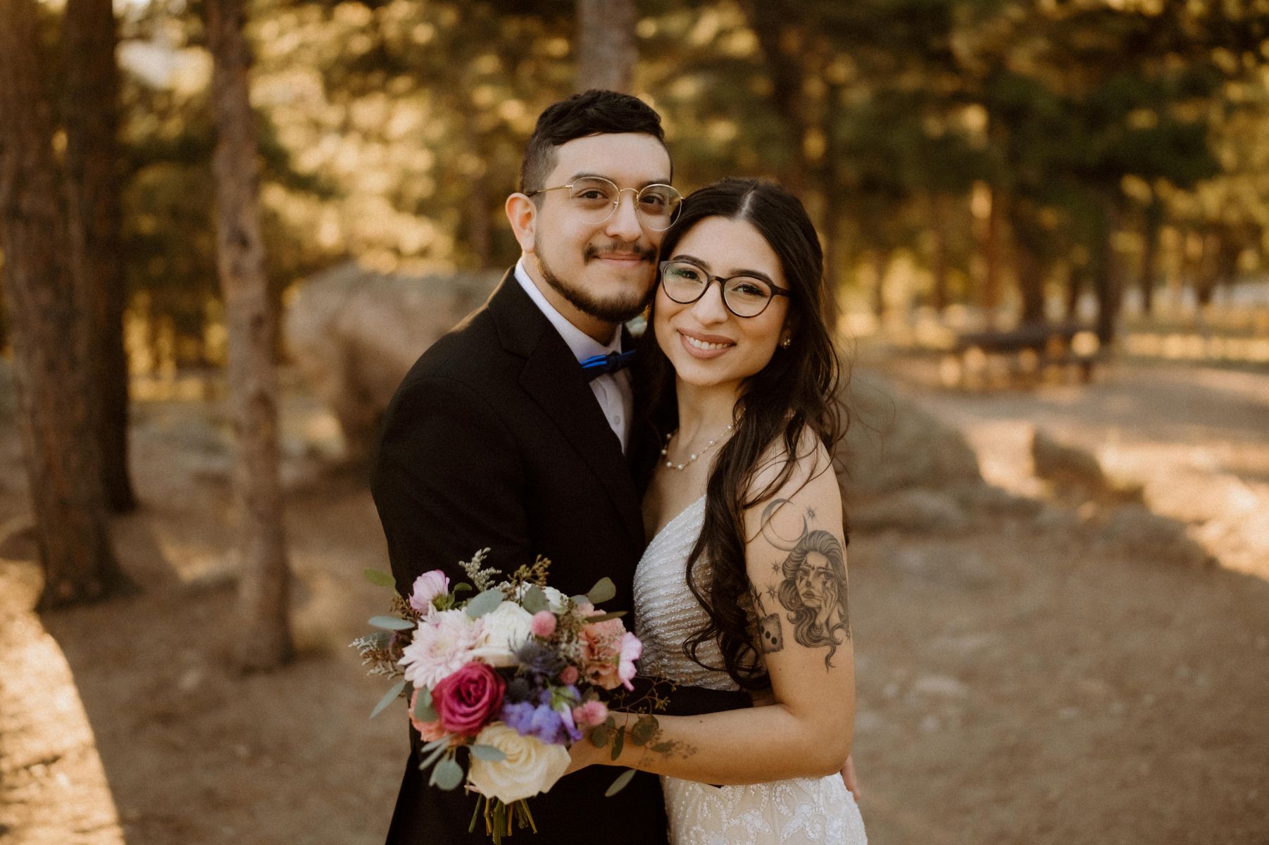 a couple in the wood embracing the woman is wearing a white wedding dress and holding a bouquet of flowers the man is wearing a black suit 