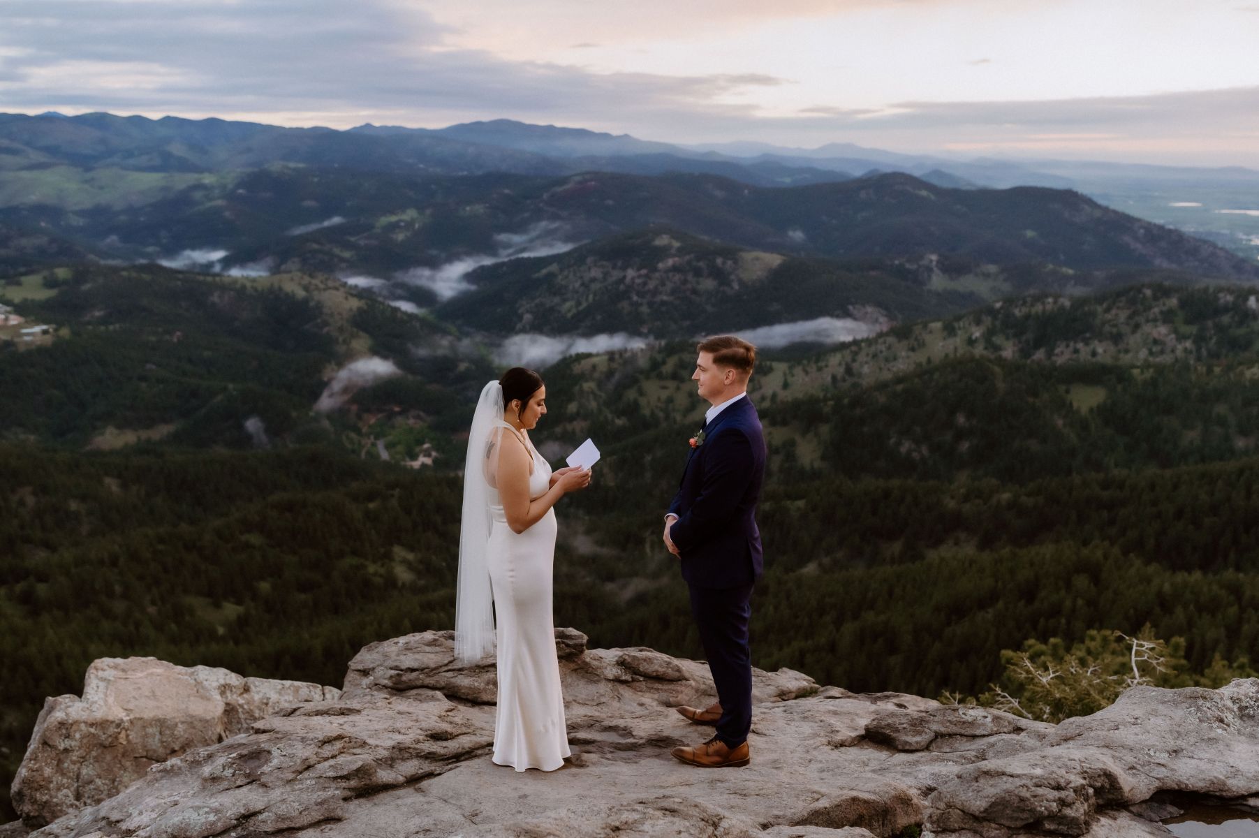 a couple having their elopement ceremony on top of an overlook next to the mountains the woman is wearing a white wedding dress and veil the man is wearing a black suit 