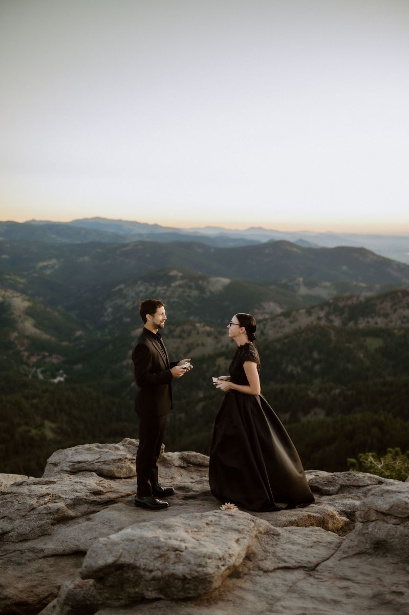 a couple reading vows to each other on top of a rocky overlook by the mountains the woman is wearing a black dress the man is wearing a black suit 