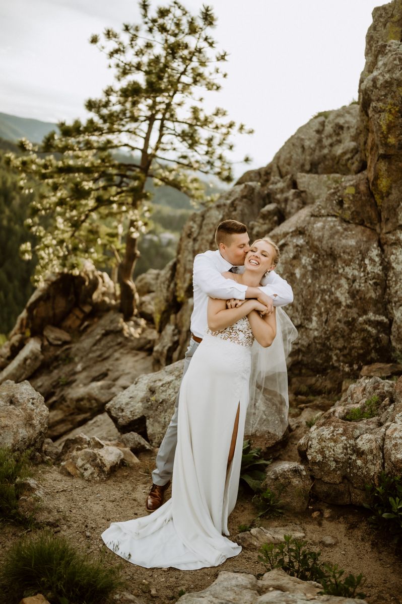 a man embracing his partner from behind on top of a rocky overlook the woman is wearing a white wedding dress the man is wearing a white button up 