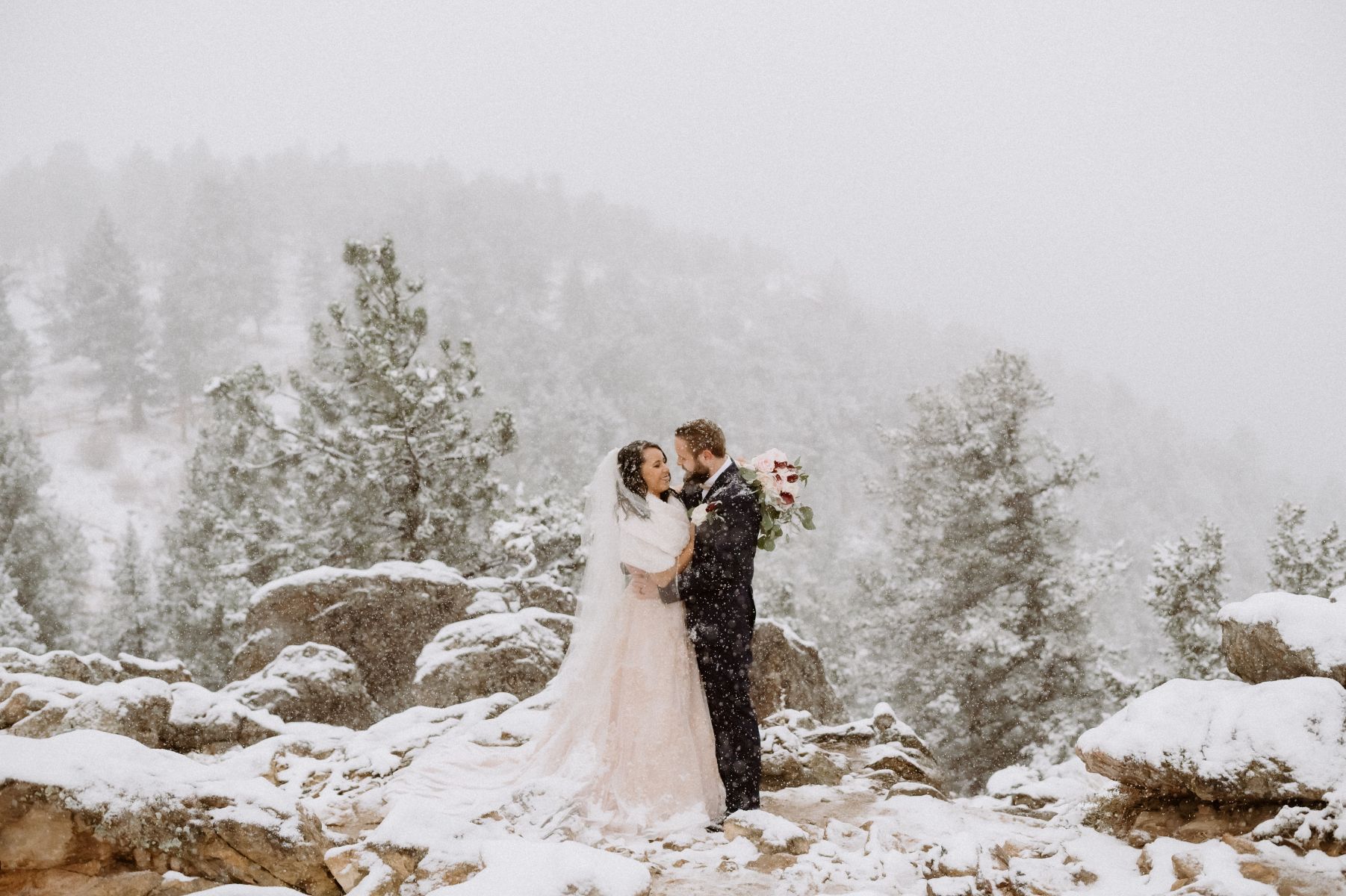 a couple having a winter elopement at lost gulch overlook in colorado it is snowing around them and they are embracing on top of rocks the woman is wearing a white wedding dress and veil the man is wearing a black suit 