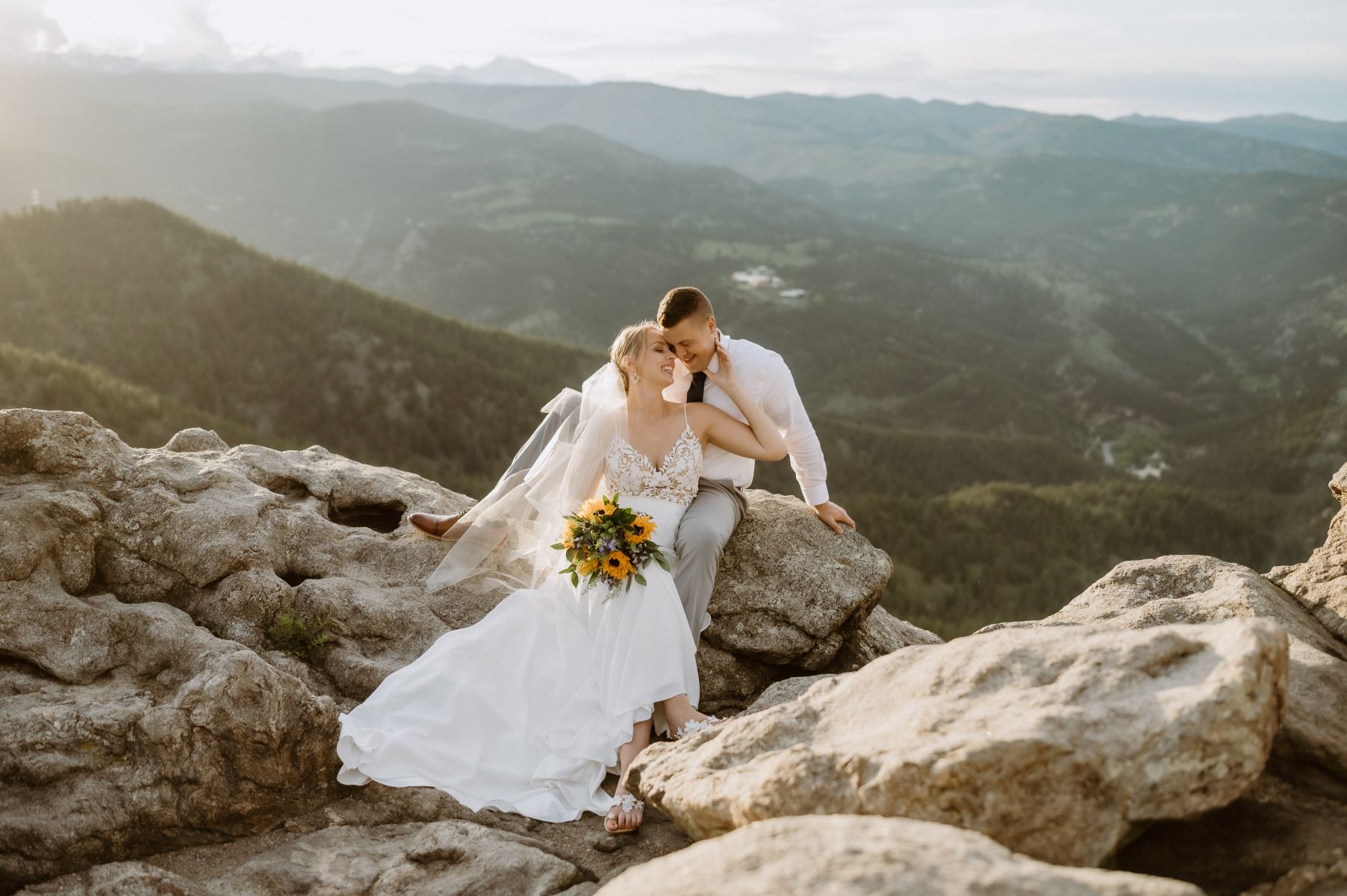 a couple sitting on top of rocks next to an overlook the woman is wearing a white wedding dress and veil and holding flowers the man is wearing a white button up 