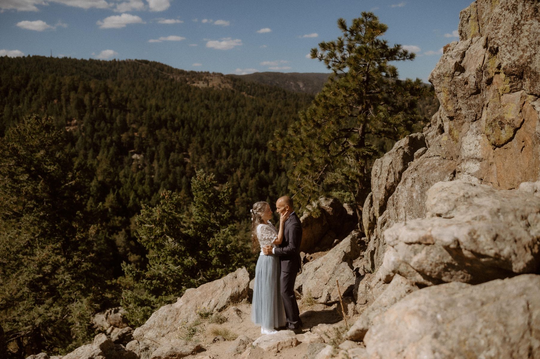 a couple on a rocky overlook in colorado behind them is a valley and pine trees the woman is wearing a white floral top and blue skirt the man is wearing a black suit and they are embracing 