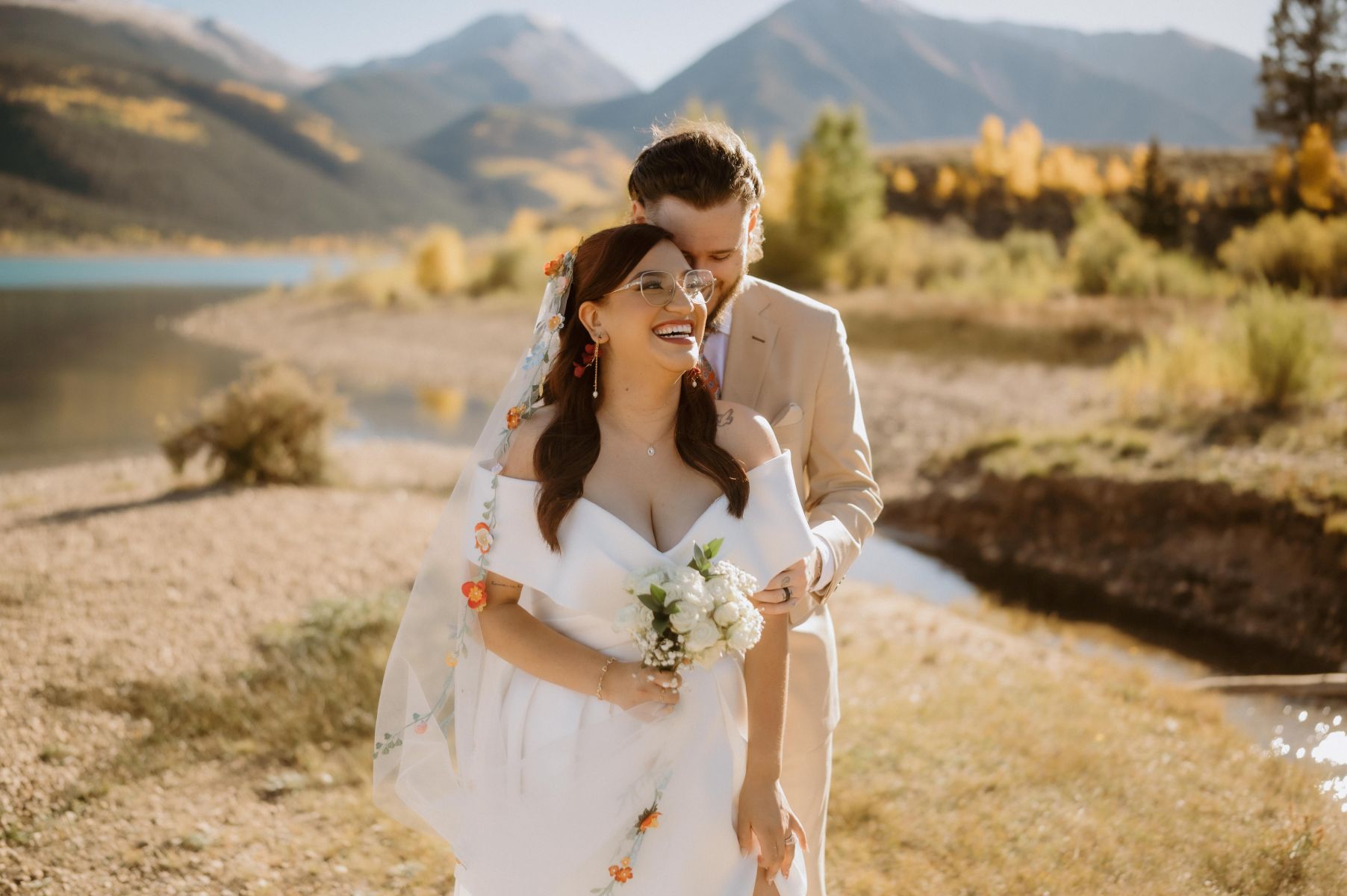 a man in a tan suit embracing his partner from behind next to a lake she is wearing a white wedding dress and holding a bouquet of flowers they are both smiling