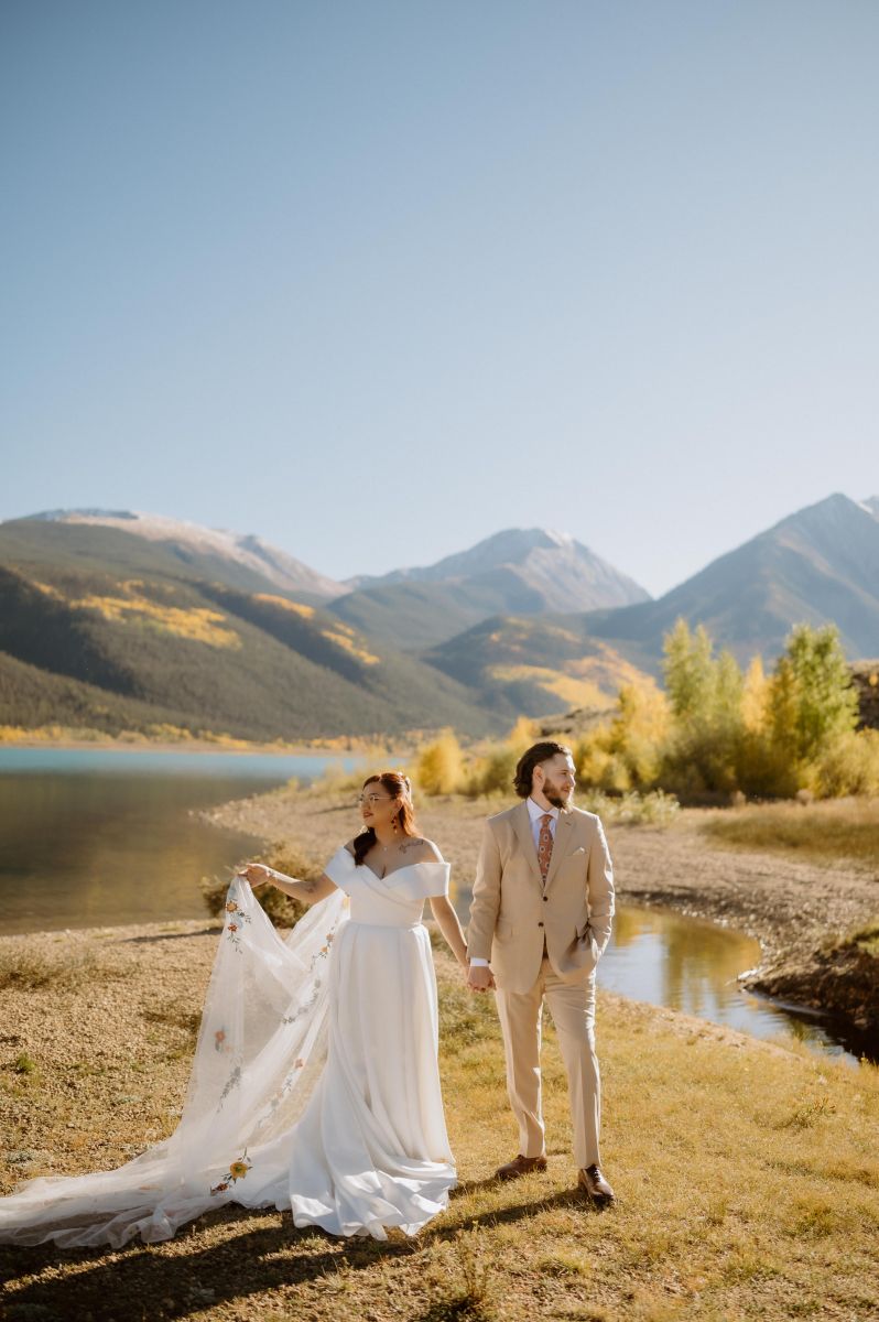 a couple taking potraits together next to a lake and the mountains at sunset the woman is wearing a white wedding dress and vei land the man is wearing a tan suit they are both holding hands 