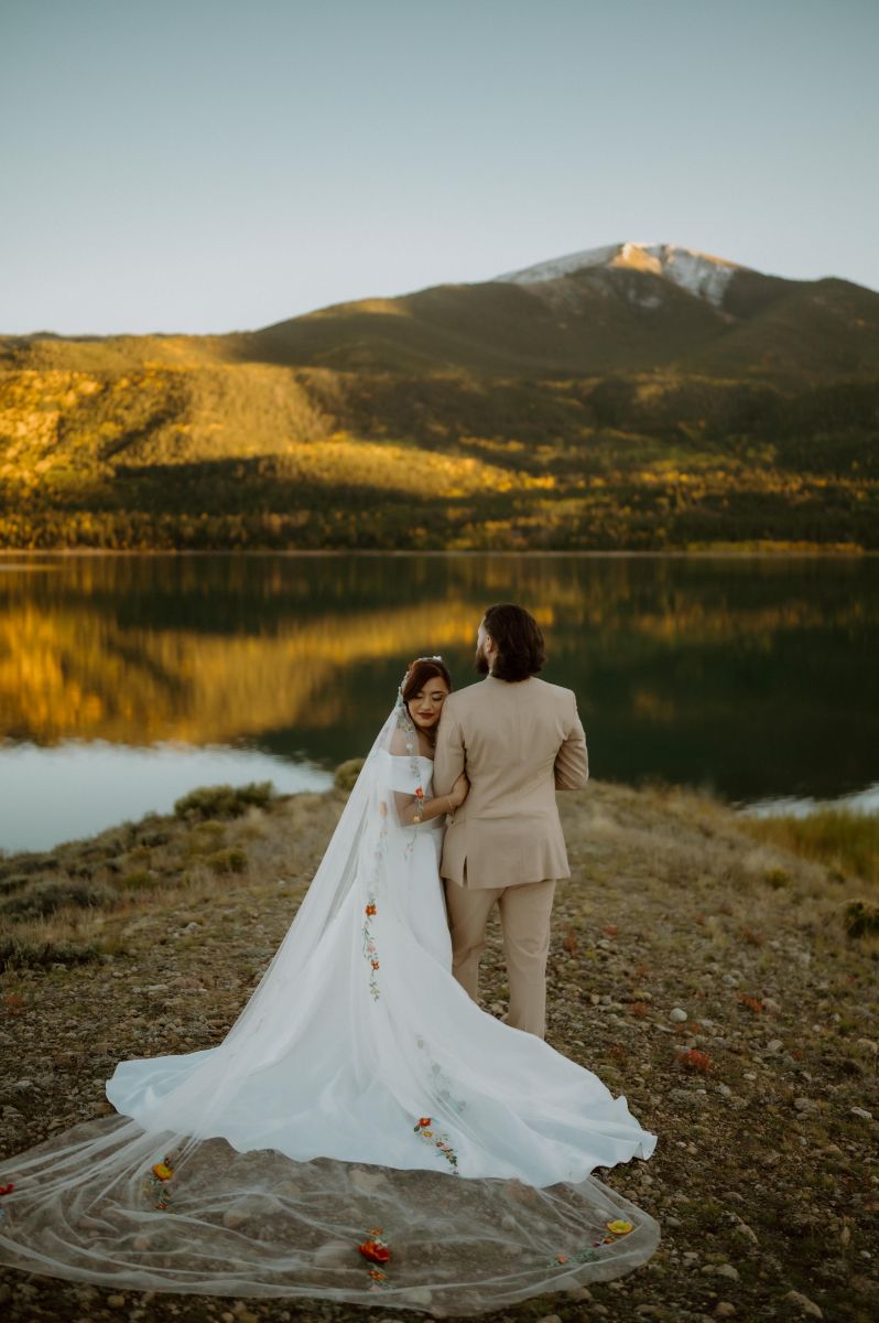 a man hugging his partner next to a lake and mountains at sunset the woman is wearing a white wedding dress and veil and hugging her partner's arm 