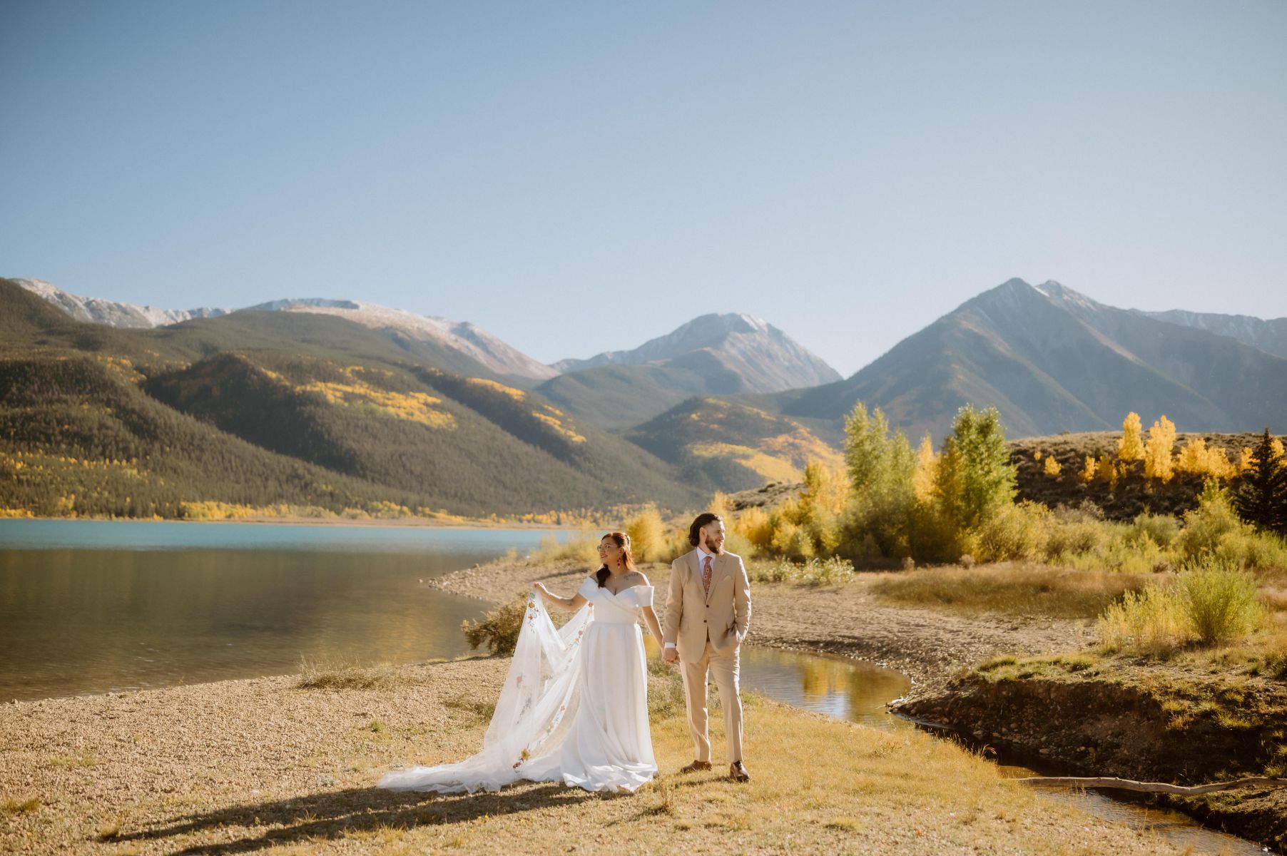 a couple standing next to a lake taking couple potraits they are surrounded by mountains the woman is wearing a white wedding dress and veil and the man is wearing a suit 