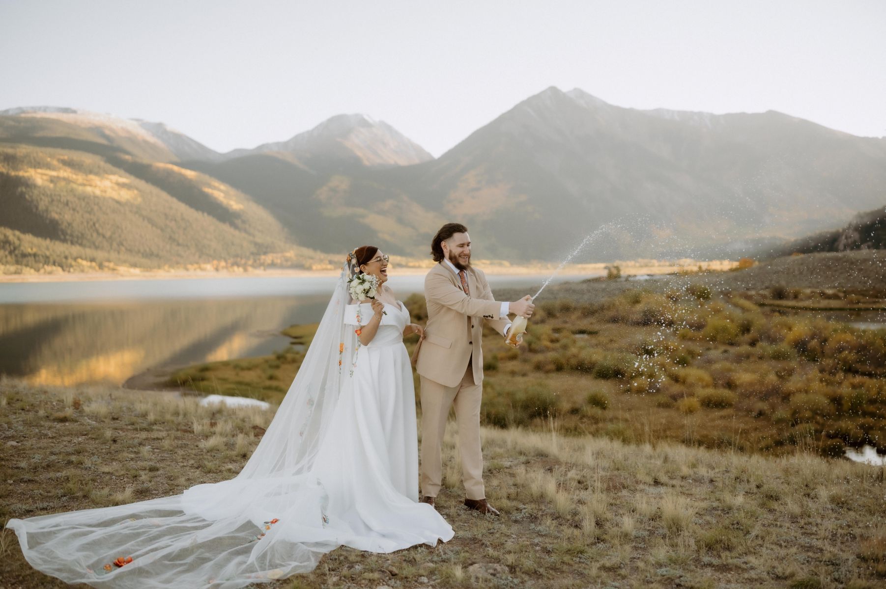 a man in a tan suit popping a bottle of champagne next to his partner who is wearing a white wedding dress and veil 