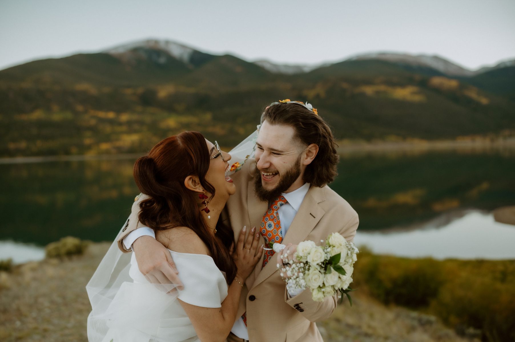 a couple embracing and laughing together next to a lake surrounded by mountains 