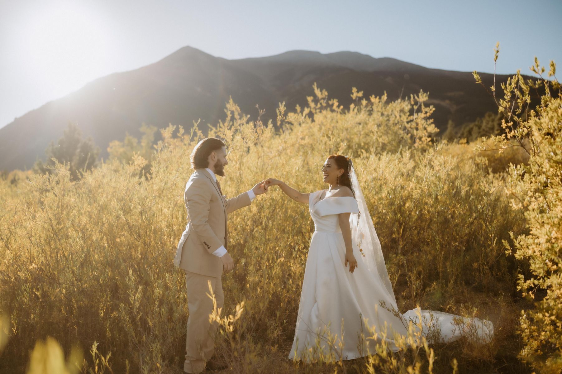 a copule dancing in the fields next to the mountains at sunset during golden hour the woman is wearing a white wedding dress and veil and the man is wearing a tan suit 