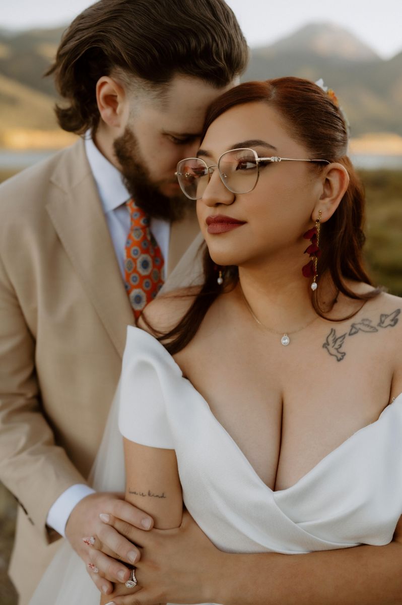 a man placing his had against his partner's face his partner is wearing glasses and looking off to the distance the man is wearing a tan suit the woman is wearing a white wedding dress
