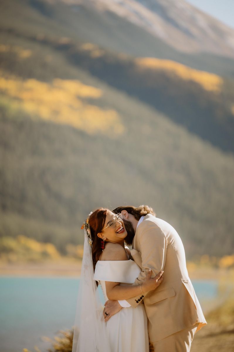 a man kissing his partner's neck she is laughing and wearing a white wedding dress and veil and he is wearing a brow suit 